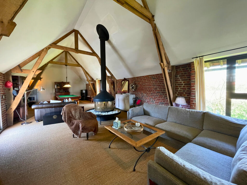 Détail des éléments anciens du salon : poutres bois, mur en briques et cheminée centrale, dans un espace chaleureux et ouvert